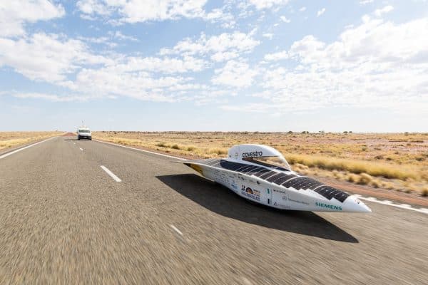 Solarbetrieben - Rennwagen "Covestro Adelie" aus Kunststoff fährt auf einer geraden Straße mit Teamwagen im Hintergrund.