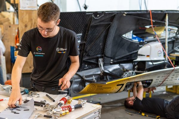 Im Vordergrund tüftelt ein Team-Mitglied des Teams Sonnenwagen Aachen am Werkzeugtisch. Im Hintergrund schraub ein Monteur auf dem Rücken liegend am solarbetrieben Rennwagen Adelie Covestro.