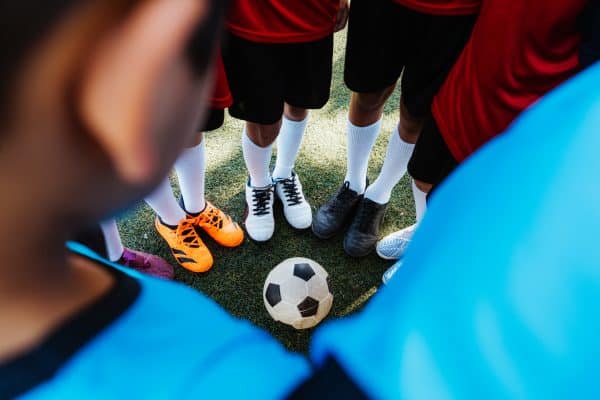 Young Soccer Players Standing Together On The Field: A close-up of young soccer players' feet in a circle around a soccer ball on the turf.