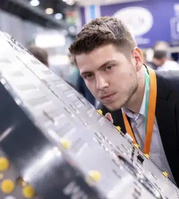 Momentaufnahme des Messestandes des GKV Auf der K 2025. Junger Mann schaut sich Industriewerkstück aus der Nähe an.
